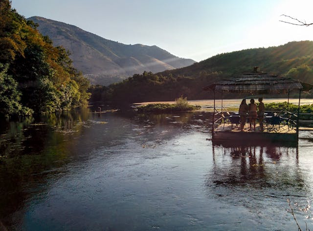 People on a water hut, river, trees, sunshine, hills, and forests.